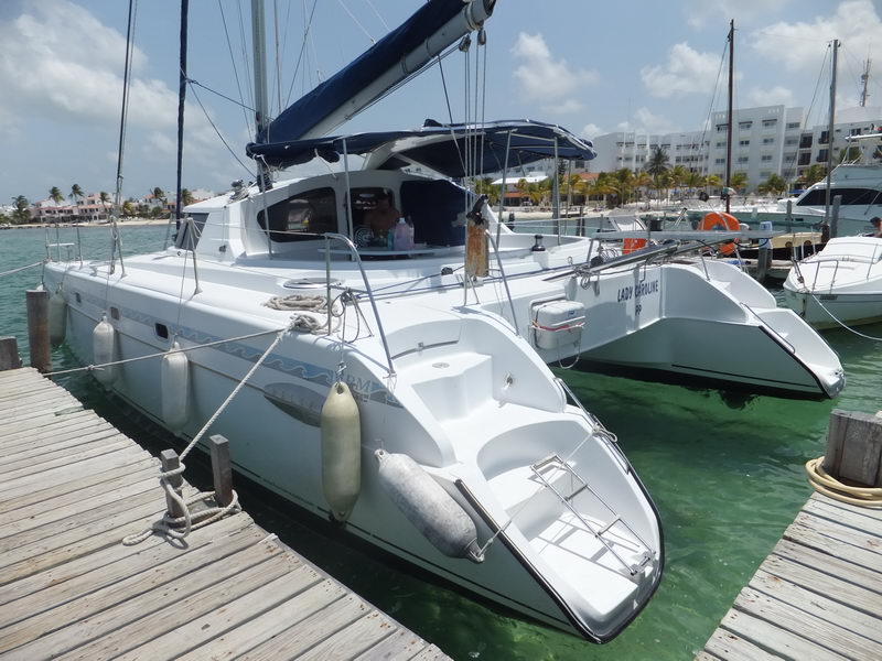 Stern quarter view of a white sailing catamaran named 'Lady Caroline' moored at a dock, twin hulls fitted with white fenders. Other boats and resort buildings behind, turquoise water, sunny.