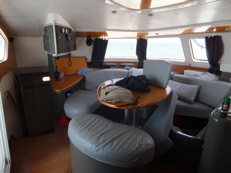Interior salon of a catamaran with a grey leather wraparound settee and varnished wood table, a navigation/instrument panel on the bulkhead and sea visible through the windows. Casual clutter - jacket, bug spray - on the table.