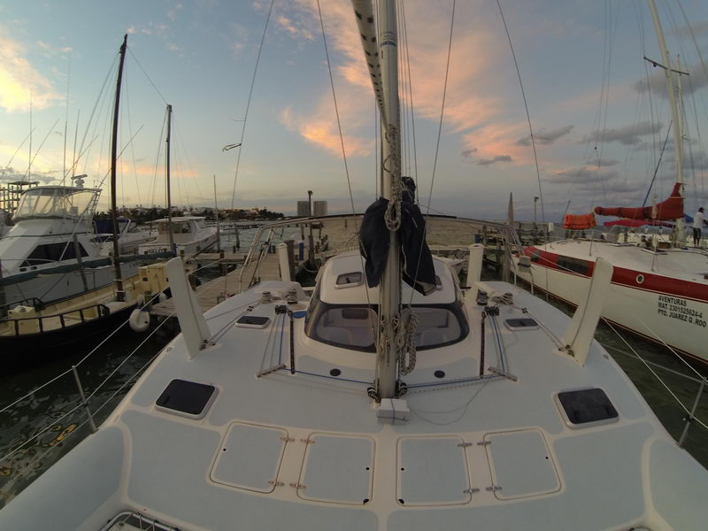 Wide-angle bow view from the deck of a catamaran at a marina at sunset, mast and furled sail centered. Pink-tinged clouds overhead; sportfishing boats and a red-hulled boat moored nearby.