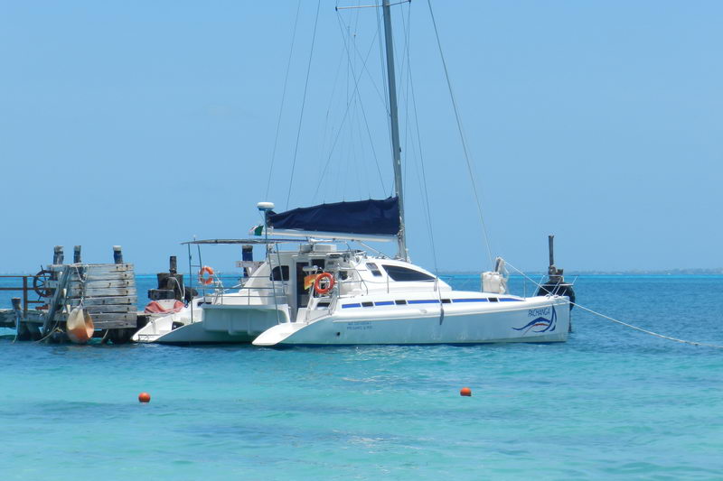 White sailing catamaran 'Pachanga' moored off a small wooden dock in shallow turquoise water, blue bimini and mooring buoys floating nearby. Bright sunny day, calm flat sea.