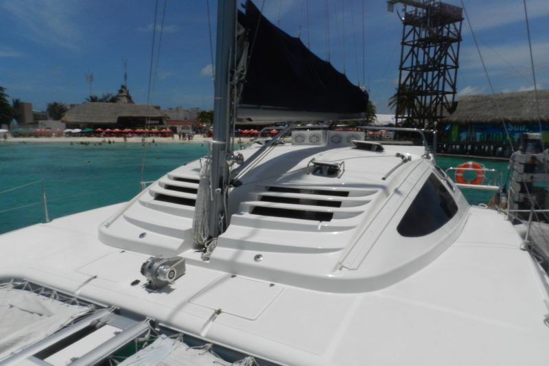 Foredeck view of a sailing catamaran looking toward the cabin coachroof and a furled black sail, anchored off a beach lined with palapas, red beach umbrellas and an observation tower onshore, sunny.
