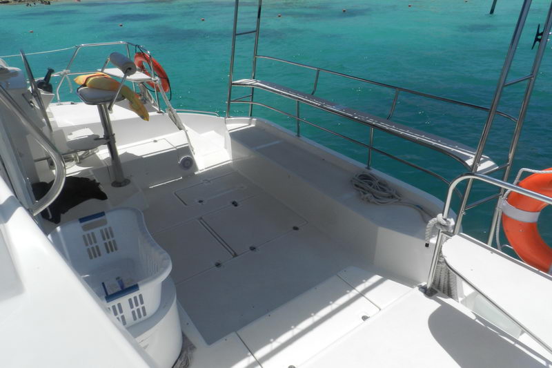 Aft deck and cockpit of a catamaran looking over the stern to turquoise water, with stainless rails, a helm seat with colorful cushion, a coiled rope, a laundry basket and an orange life ring. Bright sun.