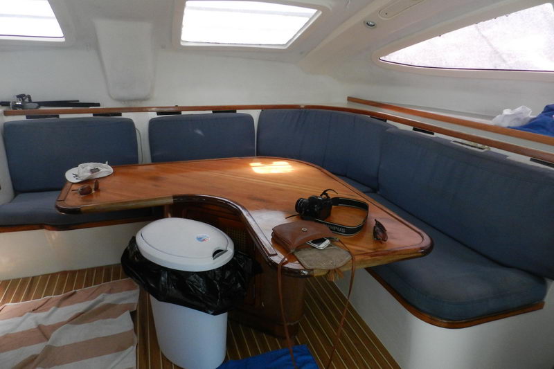 Interior salon of a sailing catamaran with a U-shaped settee in grey-blue cushions, a varnished wood table, a trash bin underneath and a camera and sunglasses on the table. Wood-plank cabin sole.