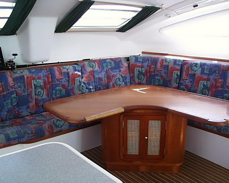 Interior salon with a U-shaped settee upholstered in 1990s-style abstract blue, red and green patterned fabric, a varnished wood table with inlay over a cane-door cabinet base and green curtains above.