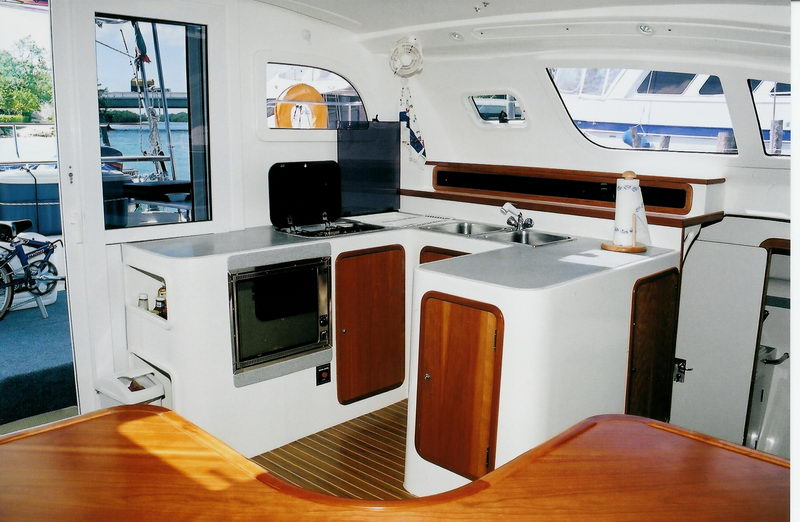 Interior galley of a catamaran with white counters and a sink, a two-burner stove, a microwave/oven and wood cabinet doors. A window looks out onto the marina and moored boats.