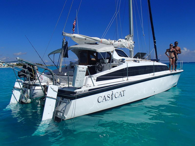 Small black-and-white hulled 'Casi Cat' sailing catamaran (a MacGregor-style cat) anchored in brilliant turquoise water, with a couple posing on the side deck. Furled mainsail, hardtop bimini, twin outboards and dive gear at the stern; US flag flying. Bright blue sky.