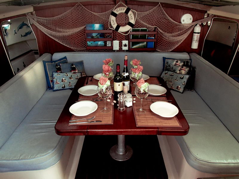 Overhead wide view of a sailing yacht's salon dining table set for a meal: varnished wood table with white plates, oversized wine glasses, a wine bottle and pink napkin roses, surrounded by light-blue cushioned settees with lighthouse-print pillows. A decorative fishing net and 'EAT SLEEP SAIL' life ring hang on the wood bulkhead.