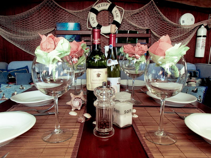 Close-up of a yacht's set dinner table with two oversized wine glasses holding pink-and-green napkin roses, two wine bottles, a glass pepper grinder and salt shakers on a bamboo runner, with seashells scattered around. The 'EAT SLEEP SAIL' life ring and fishing net blur in the background.