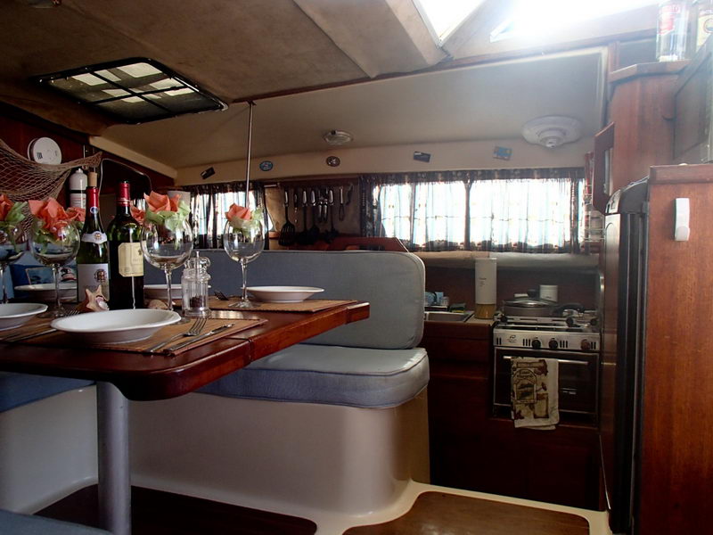 Interior of the catamaran looking aft toward the galley: a varnished wood dining table set with plates and wine glasses on light-blue settees in the foreground, with a gimballed stove/oven, hanging kitchen utensils, curtained ports and a fridge along the side. Warm wood tones and daylight through the windows.