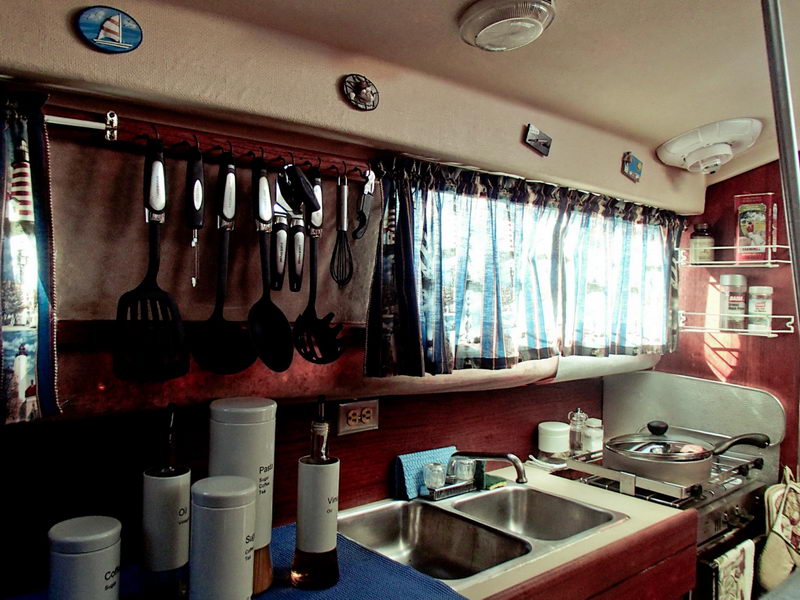 Compact yacht galley with a double stainless sink, two-burner stove and small pan, kitchen utensils hanging from a rail, white storage canisters and spice jars, and curtained windows. Warm reddish wood tones and nautical decals throughout.