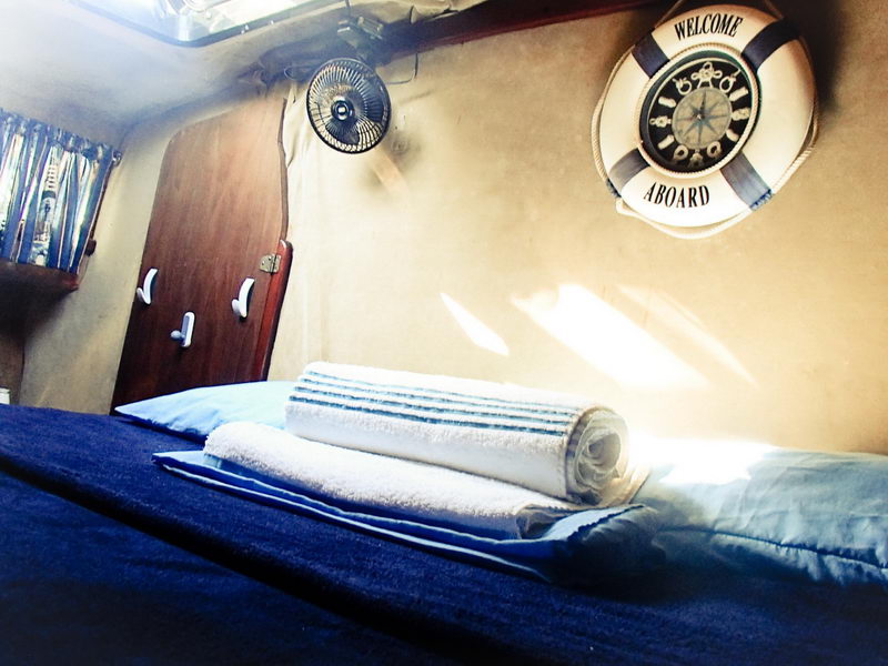V-berth cabin of a sailing yacht with a navy-covered double bed, white pillow, rolled white-and-blue striped towels, a small oscillating fan and a 'WELCOME ABOARD' compass-design life ring on the tan bulkhead. Low-angle warm-toned shot.