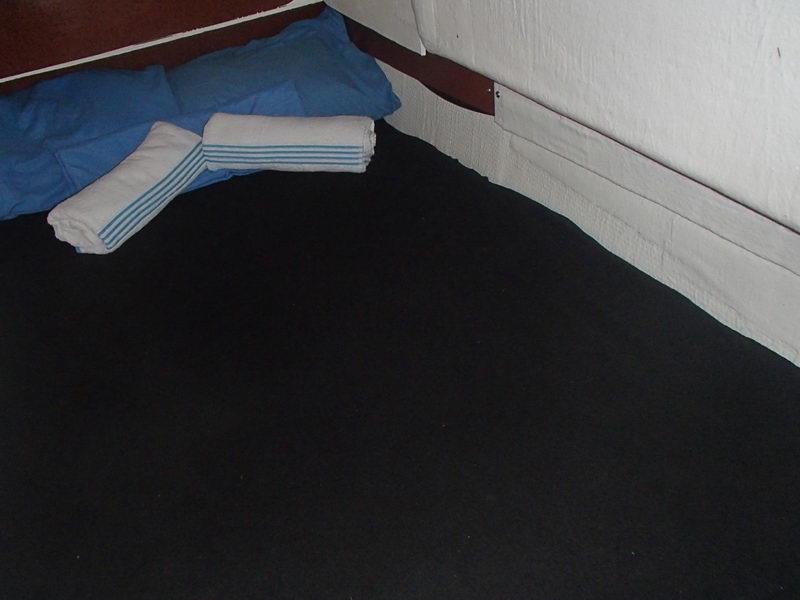 Close-up of the corner of a yacht berth showing a dark navy mattress cover, a blue pillow and a rolled white towel with blue stripes against the white hull liner. Plain, slightly dim documentary-style shot.