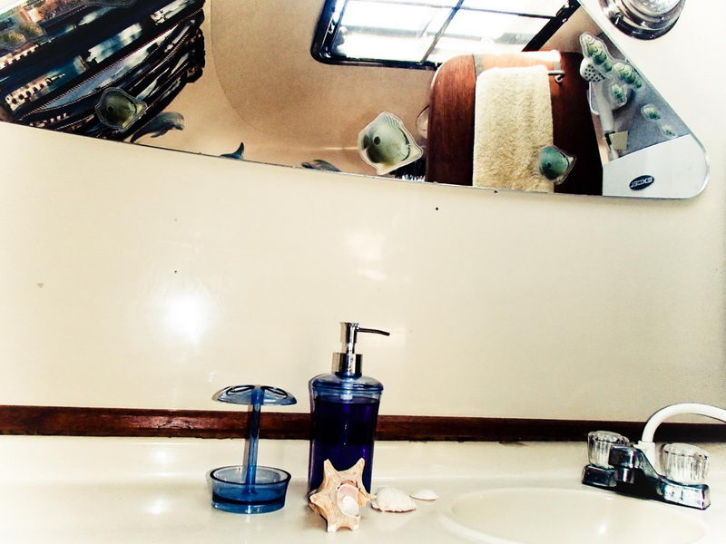 Yacht bathroom vanity with a white sink, chrome faucet, blue soap dispenser and toothbrush holder, seashells and a starfish on the counter. An angled mirror above reflects a towel and more fish decals.
