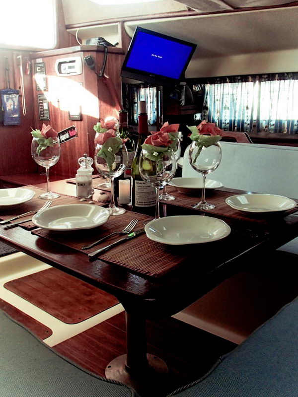 Interior salon of a sailing yacht set for dinner, viewed vertically: varnished wood table with white plates, wine glasses and napkin roses, a small flat-screen monitor switched on above, and instrument panels and curtained ports on the wood bulkhead.