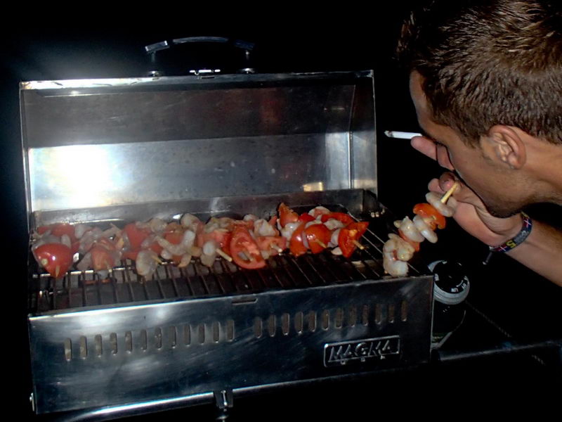 Nighttime shot of a man tending shrimp-and-tomato skewers on a stainless Magma marine grill, leaning in to inspect the food. Dark background with the grill brightly lit.