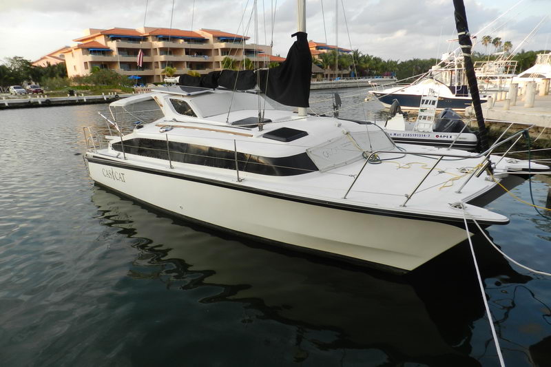 Black-and-white hulled small sailing catamaran 'Casi Cat' (a MacGregor-style cat) moored bow-out in a marina, with a furled mainsail in a black cover and low cabin top. Calm dark water reflects the hull; a tan resort condo building, palms and other boats sit behind under an overcast sky.