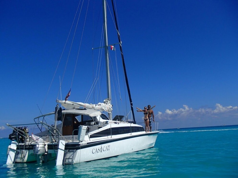 Small black-and-white sailing catamaran 'Casi Cat' anchored in brilliant turquoise water with a couple posing on the forward deck. Mast with furled sail, outboard and dive gear on the stern; deep blue sky overhead.