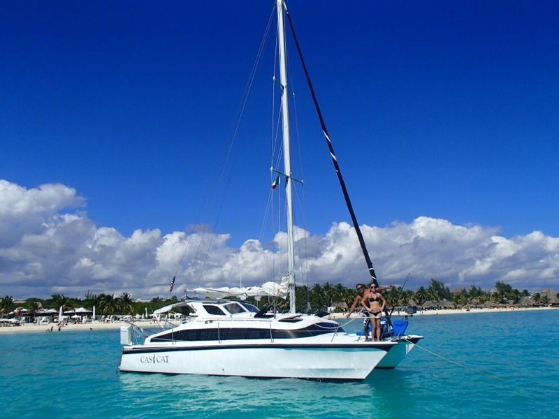 The 'Casi Cat' catamaran anchored in turquoise shallows off a palm-lined beach, a couple standing on the bow. Tall mast and boom dominate the frame against big white cumulus clouds and blue sky.