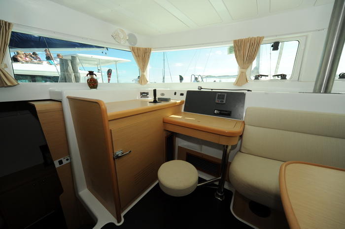 Interior of a catamaran showing the navigation/chart table with a folding instrument panel and a round stool, light wood cabinetry, a cream settee and panoramic curtained windows looking onto the marina.
