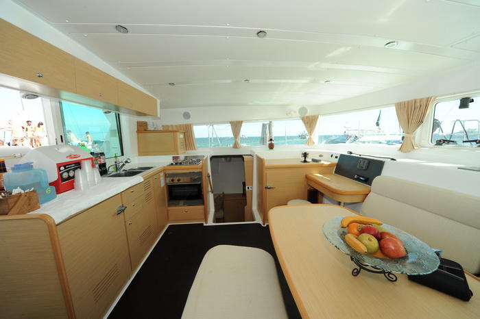 Interior galley and salon of a Lagoon catamaran with a stainless sink, gas stove and oven, light wood cabinetry, a dining table with a bowl of fruit, panoramic windows and dark flooring.