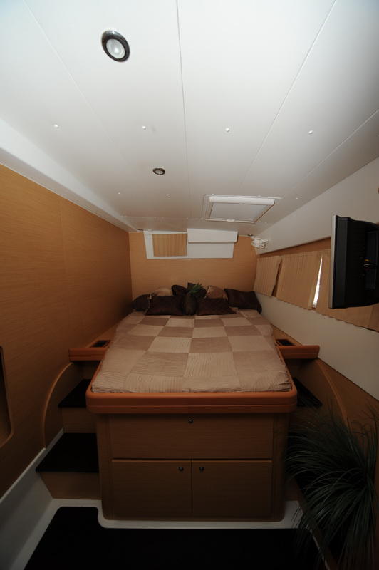 Interior double cabin of a catamaran with a queen bed dressed in a beige checkered cover and brown cushions, wood paneling, a wall-mounted flat TV, overhead hatch and recessed lighting.