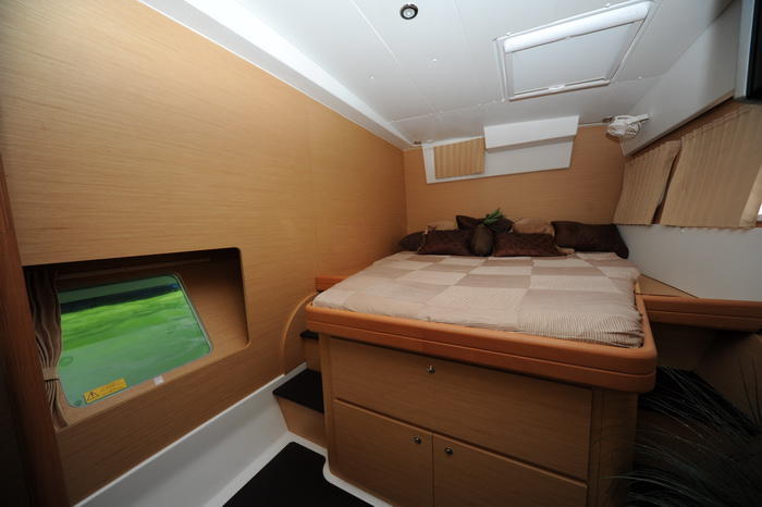 Interior double cabin of a catamaran with a beige checkered bed and brown pillows, light wood paneling, a hull-side window showing green water outside, steps and storage drawers.
