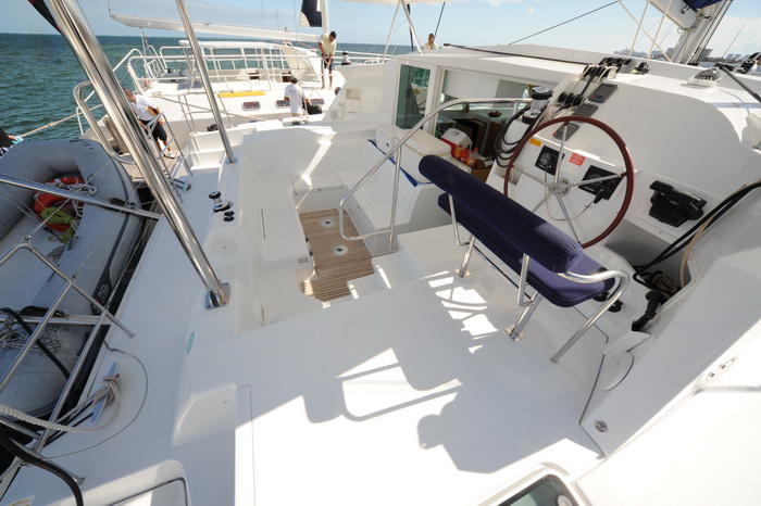 View across the bright white deck of a Lagoon catamaran looking forward from the cockpit, showing the cushioned helm seat and wheel, a dinghy stowed on davits, a couple of crew and open sea on a sunny day.