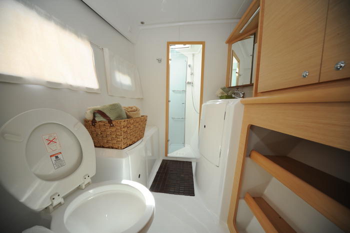 Interior head of a catamaran with a white marine toilet, a basket of rolled towels, a glass-door shower stall, a sink and mirror, and light wood cabinetry and shelving.