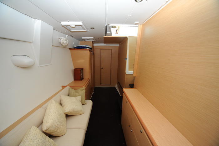Interior hull passageway of a catamaran with a cream settee and throw pillows, light wood paneling and cabinetry lining a narrow corridor leading to cabin doors over a dark floor.
