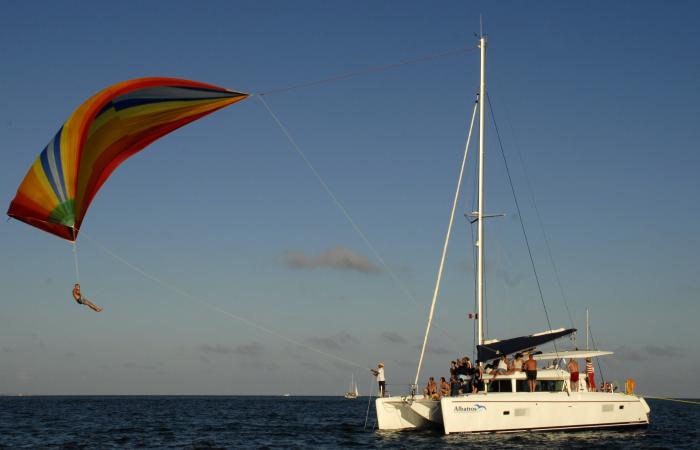 A sailing catamaran named 'Albatros' at dusk with passengers aboard while a person hangs from a colorful rainbow parasail-style sail flown off the side, calm sea and warm evening light.