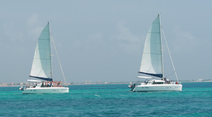 Two white sailing catamarans with navy-trimmed sails cruising side by side over turquoise Caribbean water with passengers aboard, a hazy coastline with buildings in the distance.