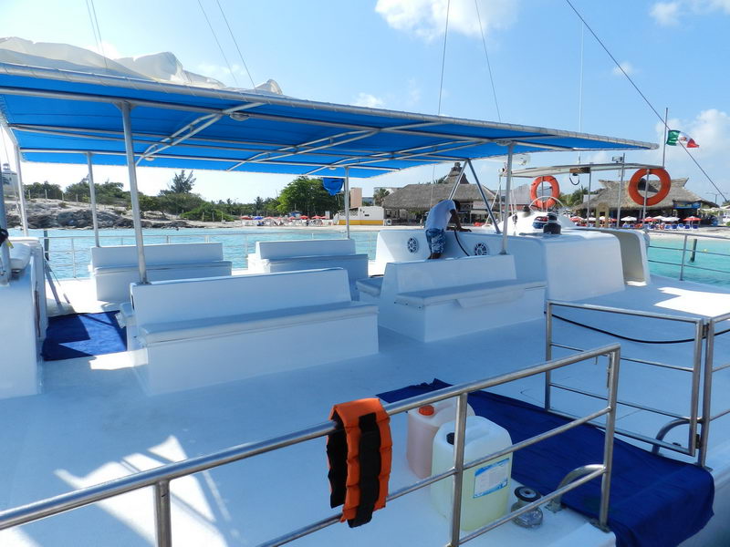 Upper passenger deck of a large catamaran with rows of molded white benches under a blue canvas top, life rings and a crew member, with a beach of palapas and sunbathers visible behind.