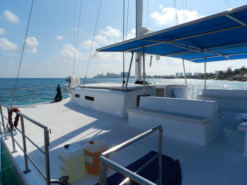 Wide foredeck of a large catamaran with bench seating and a blue bimini, looking out over calm sea toward the Cancun hotel-zone skyline on a sunny day, water jugs on deck.