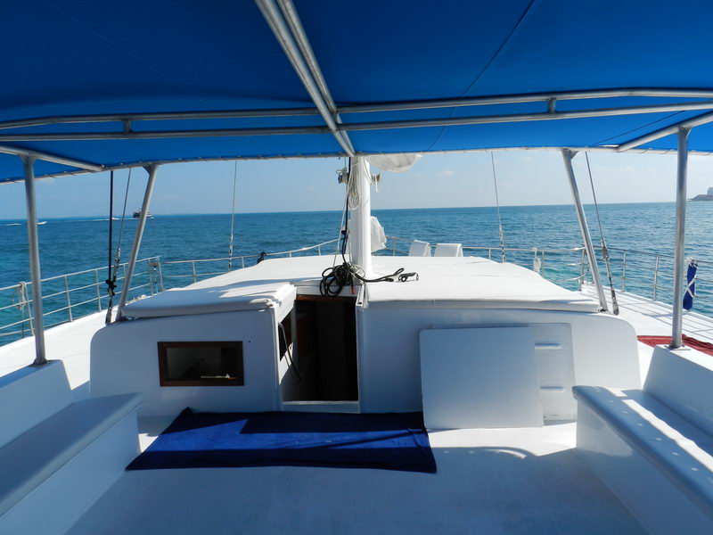 View from under the blue canvas bimini of a catamaran looking forward across white deck cushions and benches toward open blue sea, rigging and a rolled sail overhead.