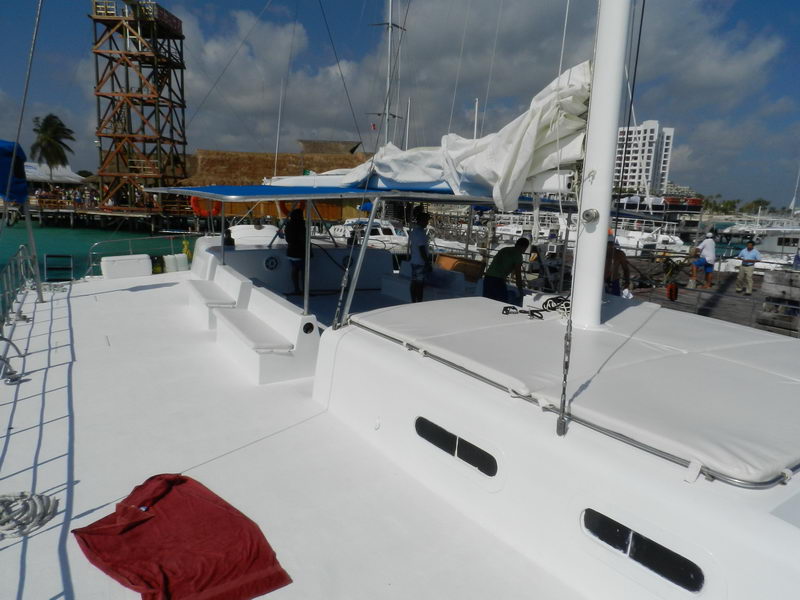 Foredeck of a large catamaran with white deck cushions and a red shirt left out, crew working near the mast, a marina with a tall wooden tower, other boats and buildings behind under a sunny sky.
