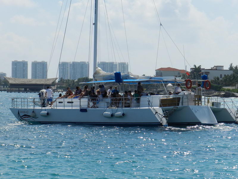 Side view of a large party catamaran named 'Samba 3' crowded with passengers, twin hulls and a rolled sail, Cancun high-rise hotels in the background across blue water.