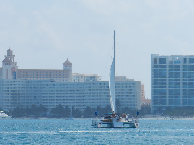 A sailing catamaran with a single white sail seen at a distance crossing in front of the Cancun hotel-zone high-rises, hazy sky and pale blue water.