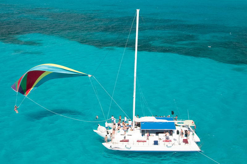 Aerial view of a large party catamaran on turquoise reef water with passengers aboard and a person suspended from a colorful rainbow parasail sail flown off the side, a blue sunpad on deck.
