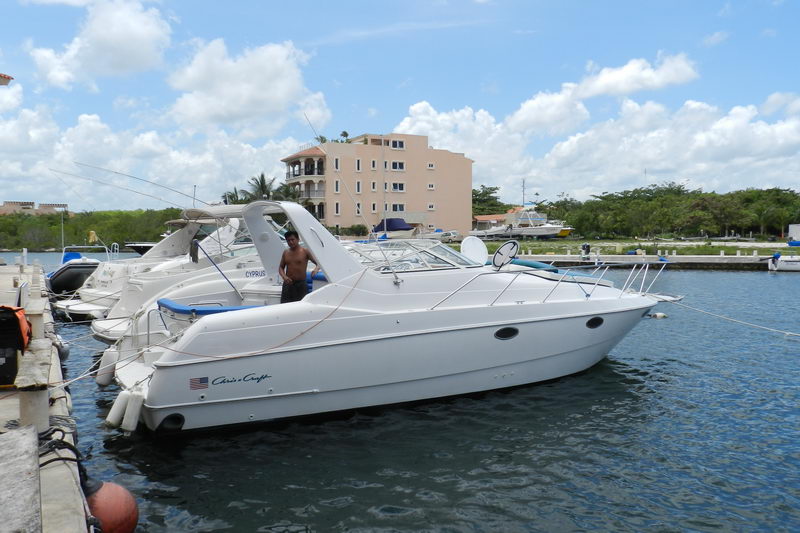 White Chris-Craft cabin cruiser named Cyprus docked at a marina, side profile view, with a US flag decal on the hull. A man stands in the cockpit; a multi-story building and trees in the background. Sunny with clouds.