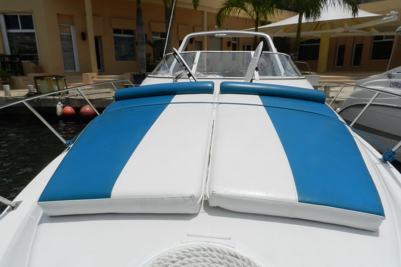 Close-up of the bow sunpad of a cabin cruiser with blue-and-white striped cushions, photographed dockside. A second similar boat and a waterfront building with umbrellas behind. Bright midday sun.