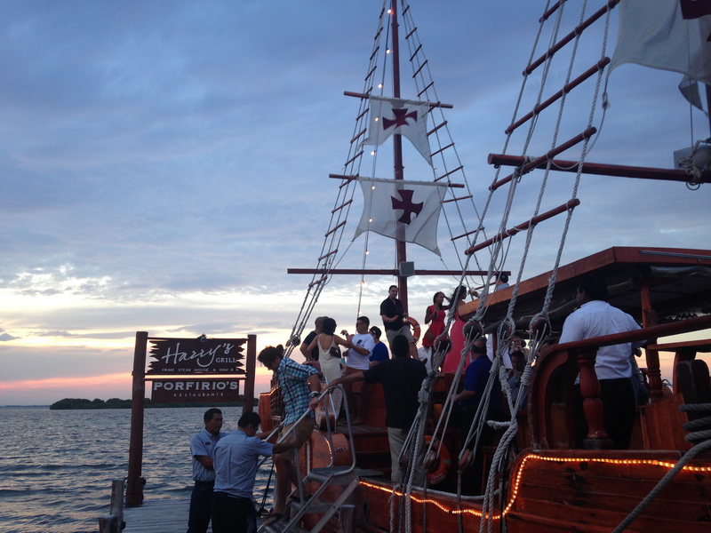 Guests boarding the Columbus galleon at dusk beside the Harry's and Porfirio's pier with cross sails and string lights glowing over the deck