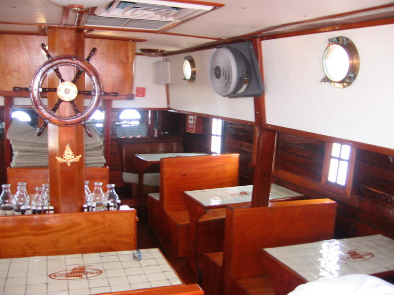 Varnished hardwood interior cabin of the Columbus galleon with the ship's wheel, tiled dinner tables and brass porthole windows