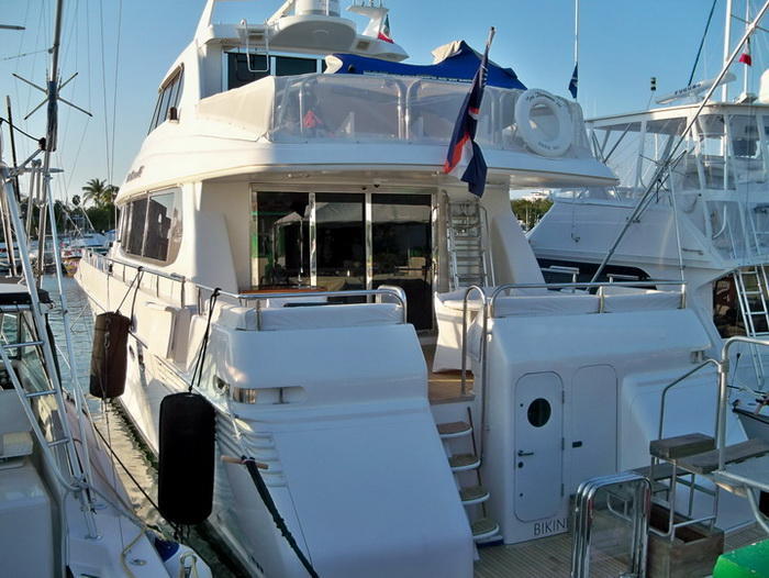 Stern view of a large white motor yacht docked at a marina, showing the cushioned aft deck, boarding stairs, swim platform and flags. Other sport-fishing boats are moored alongside in warm afternoon light.