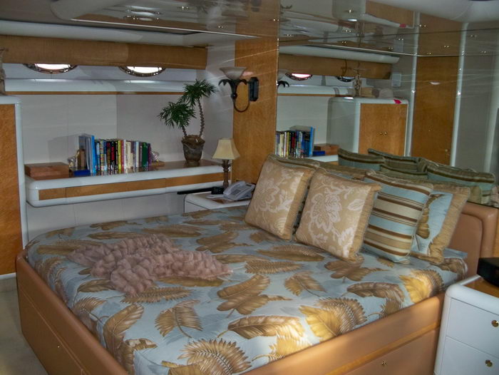 Master stateroom aboard a yacht with a large bed dressed in a gold-and-blue floral feather-print bedspread and decorative pillows. Wood cabinetry, a bookshelf with novels, a lamp and mirrored surfaces surround the cabin.