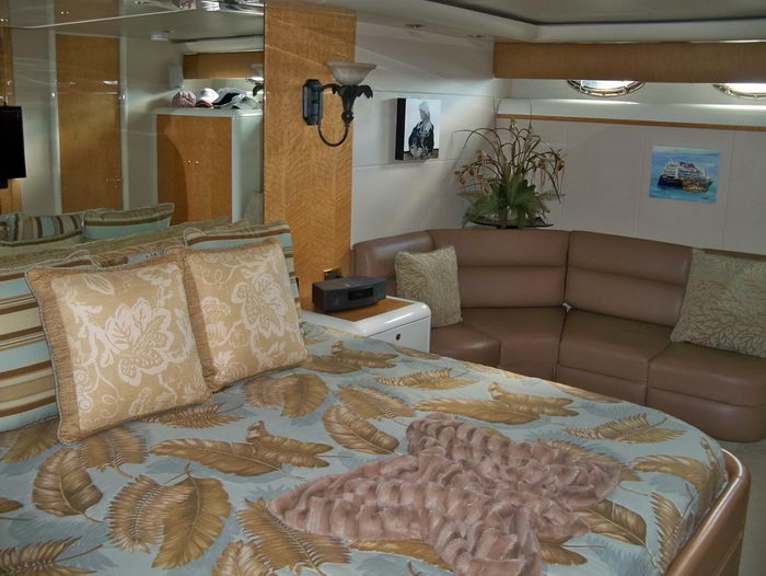 Yacht master stateroom showing a bed with a gold-and-blue feather-print bedspread and a soft pink throw, beside a brown leather corner sofa. Floral arrangements, framed art and a mounted TV decorate the warm wood-toned cabin.