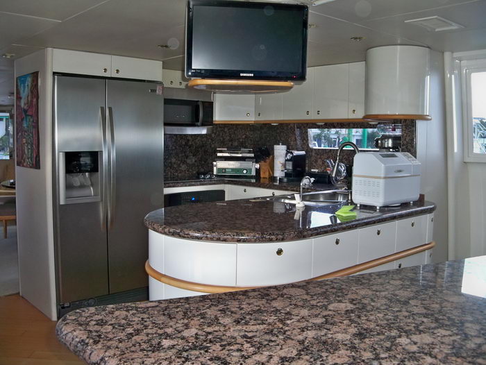 Yacht galley with a large stainless steel side-by-side refrigerator, brown granite countertops, a stainless sink, microwave, coffee and bread machines, white cabinetry and a wall-mounted Samsung TV. Bright and well-equipped.