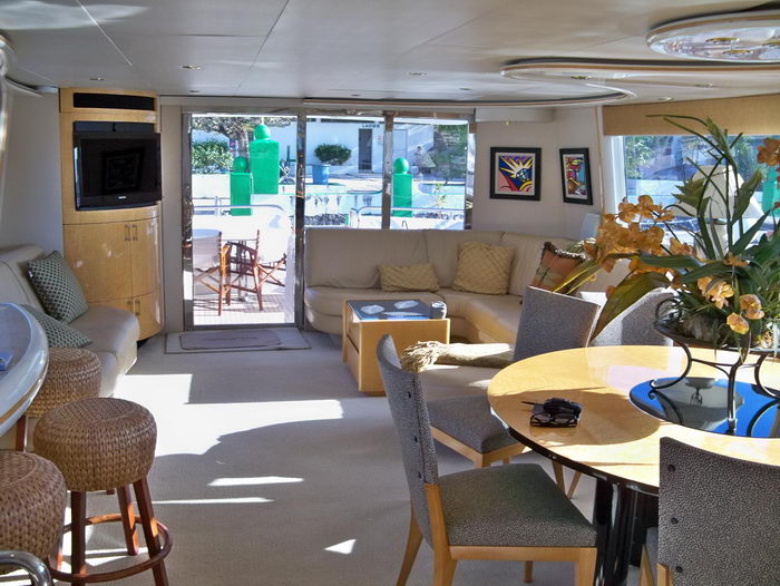 Bright yacht salon with a cream sectional sofa, a round dining table with upholstered grey chairs and woven bar stools, a corner TV cabinet and sliding glass doors opening to a sunny aft deck with a marina behind.
