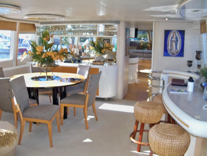 Main salon of a yacht with a round black dining table ringed by grey upholstered chairs, twin orange-flower arrangements, a curved granite bar with woven stools and a framed Virgin of Guadalupe image on the wall. Sunlit and open.