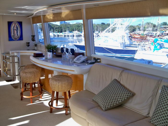 Yacht salon bar area with a curved counter, two woven bar stools, glassware and a telephone, beside a cream leather sofa with patterned pillows. Large windows reveal a marina packed with sport-fishing boats in bright sunlight.