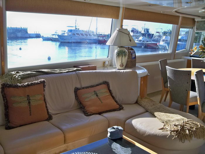 Yacht salon with a long cream leather sofa accented by orange dragonfly-print pillows and a knit throw, set beneath wide windows looking onto a calm marina with moored boats at dusk. Wood dining chairs and a table sit at right.